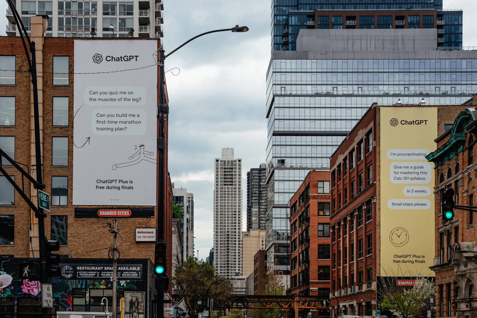 A imagem mostra uma rua urbana com edifícios altos e modernos. Dois painéis publicitários estão visíveis, um à esquerda e outro à direita, ambos com textos promocionais. O céu está nublado, e há uma mistura de arquitetura antiga e nova nas construções. No fundo, um edifício alto se destaca, possivelmente um arranha-céu.