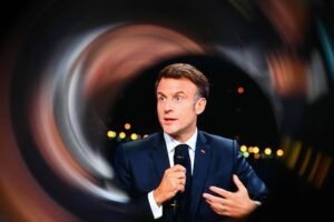 Imagem mostra Emmanuel Macron, um homem em um terno escuro e que está falando ao microfone. Ele tem cabelo castanho claro e está gesticulando com a mão esquerda. O fundo é desfocado, criando um efeito de movimento, com luzes brilhantes visíveis.