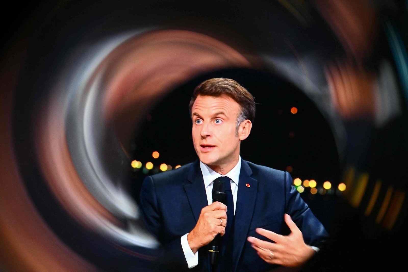 Imagem mostra Emmanuel Macron, um homem em um terno escuro e que está falando ao microfone. Ele tem cabelo castanho claro e está gesticulando com a mão esquerda. O fundo é desfocado, criando um efeito de movimento, com luzes brilhantes visíveis.