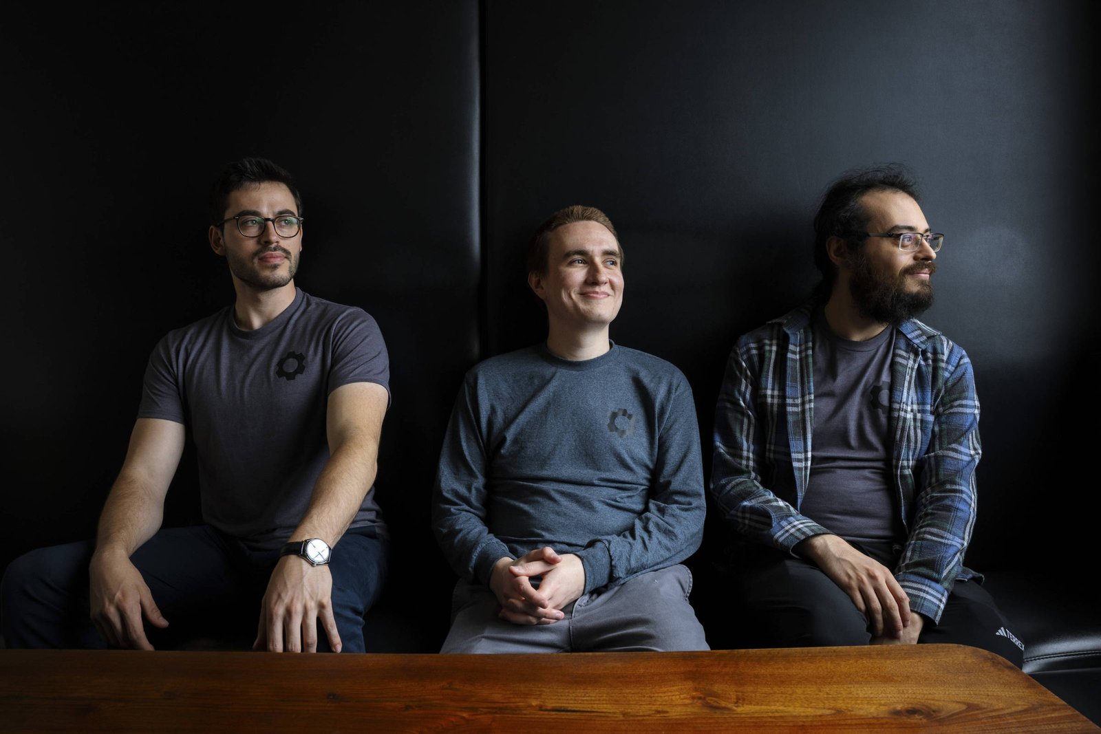 Três homens estão sentados em um sofá, com um fundo escuro. O homem à esquerda usa uma camiseta cinza e óculos, o do meio está sorrindo e veste uma camiseta azul, enquanto o da direita usa uma camisa xadrez e também óculos. Eles estão posicionados de forma alinhada, com as mãos sobre as pernas ou no colo.