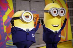 Dois personagens amarelos, conhecidos como Minions, estão vestidos com trajes formais. Um deles tem um olho e o outro tem dois olhos. Ambos estão com as mãos levantadas em um gesto de saudação ou celebração. O fundo é colorido, com elementos gráficos vibrantes.