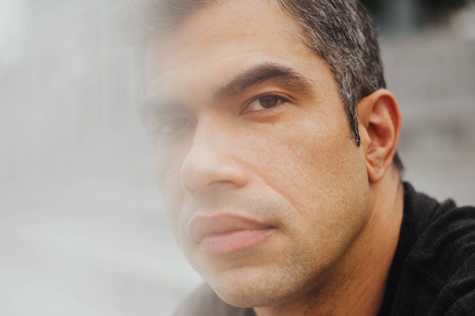 Um homem com cabelo curto e grisalho está olhando para a câmera. Ele usa uma camiseta preta e está posicionado em um ambiente urbano, com edifícios ao fundo. A imagem tem um efeito de desfoque na parte inferior, criando uma sensação de suavidade.