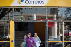 A imagem mostra a entrada de uma agência dos Correios, com um letreiro amarelo e azul. Duas mulheres estão saindo da agência. A mulher à esquerda está usando um moletom roxo e segurando um celular, enquanto a mulher à direita está vestindo uma blusa clara e segurando um papel. Ao fundo, é possível ver escadas e outras pessoas dentro da agência.