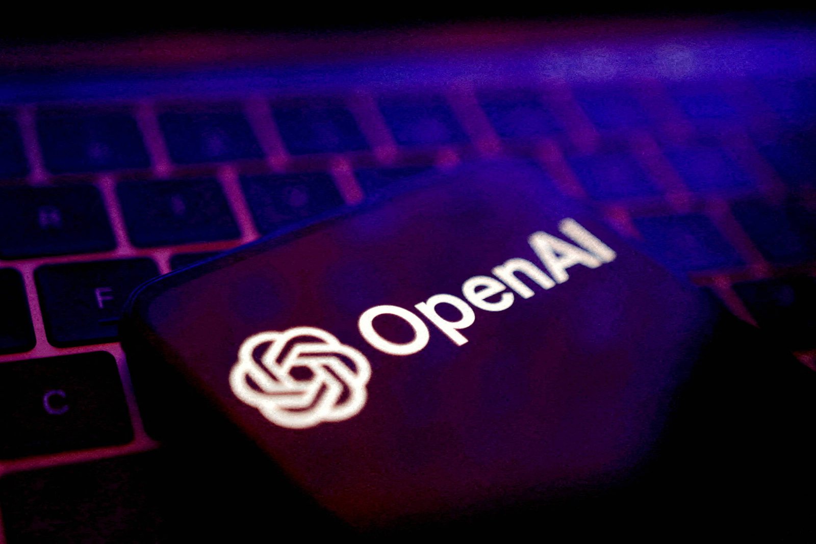 A imagem mostra o logotipo da OpenAI em um fundo escuro, sobre um teclado. O logotipo é iluminado, destacando-se em branco contra o fundo escuro e com iluminação azulada. O teclado é visível em partes, com algumas teclas em foco.