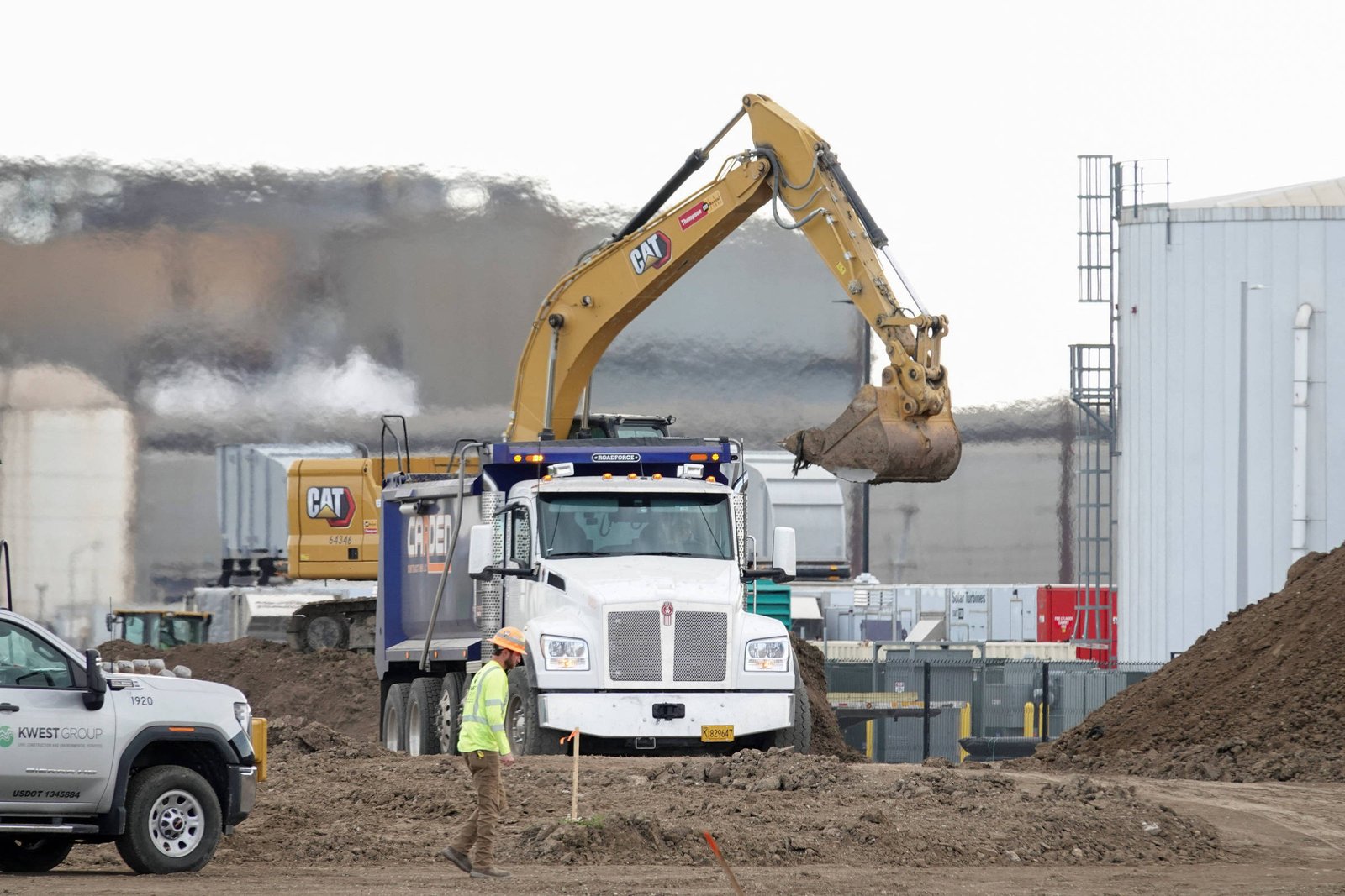 A imagem mostra um canteiro de obras com uma escavadeira amarela levantando terra. Um caminhão branco está estacionado na frente, enquanto um trabalhador vestido com colete de segurança observa. Ao fundo, há estruturas industriais e equipamentos de construção.