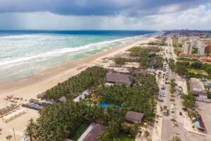 A imagem mostra uma vista aérea de uma praia com areia clara e ondas do mar. À esquerda, há várias espreguiçadeiras e guarda-sóis dispostos na areia. Na parte inferior, uma área com palmeiras e uma piscina é visível. À direita, há uma rua com edifícios e veículos, além de uma faixa de areia que se estende até o horizonte. O céu está parcialmente nublado, com algumas nuvens.