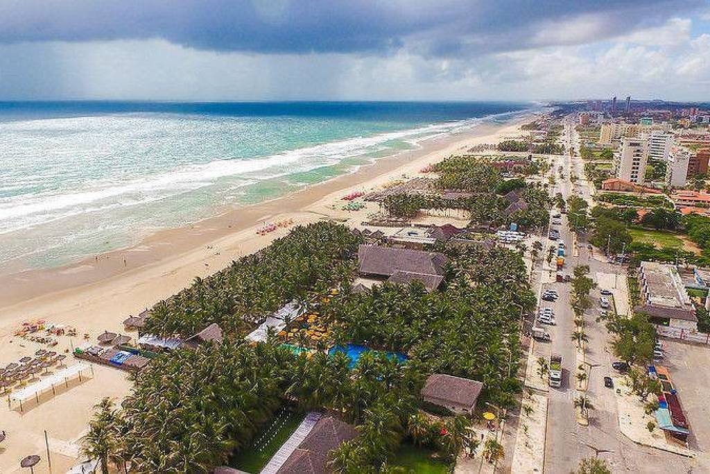 A imagem mostra uma vista aérea de uma praia com areia clara e ondas do mar. À esquerda, há várias espreguiçadeiras e guarda-sóis dispostos na areia. Na parte inferior, uma área com palmeiras e uma piscina é visível. À direita, há uma rua com edifícios e veículos, além de uma faixa de areia que se estende até o horizonte. O céu está parcialmente nublado, com algumas nuvens.