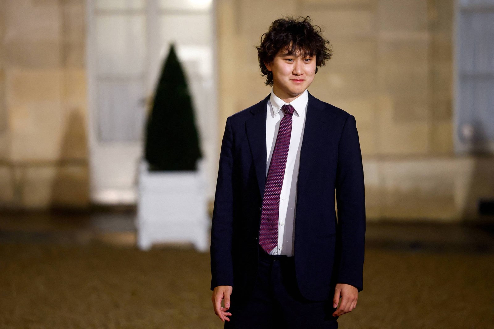 Um jovem com cabelo cacheado e bagunçado está posando em um ambiente externo. Ele usa um terno escuro, uma camisa branca e uma gravata roxa. Ao fundo, há uma planta em um vaso branco e portas de vidro. A iluminação é suave, sugerindo que a foto foi tirada à noite.