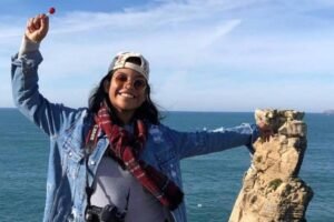 Uma mulher sorridente está em pé em um penhasco, com o mar ao fundo. Ela usa uma jaqueta jeans, uma camiseta e um boné. Em uma das mãos, ela segura um pirulito vermelho, enquanto a outra mão está levantada. A mulher tem um cachecol vermelho e preto ao redor do pescoço e uma câmera pendurada no pescoço.