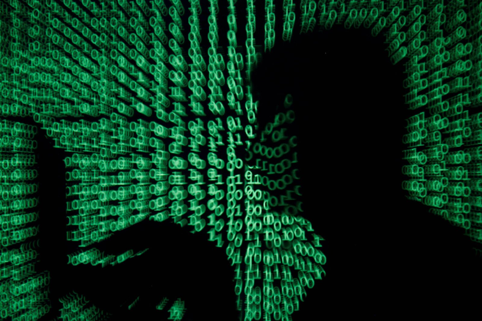 A imagem mostra a silhueta de uma pessoa em frente a um fundo repleto de números binários, representados em verde. A silhueta é escura e a iluminação do fundo destaca os números, criando um efeito visual que sugere um ambiente digital ou tecnológico.
