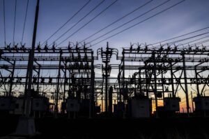 A imagem mostra uma subestação elétrica ao entardecer, com silhuetas de torres e equipamentos elétricos. O céu apresenta um gradiente de cores que vai do azul ao laranja, indicando que o sol está se pondo. Fios de alta tensão estão visíveis, cruzando a imagem.