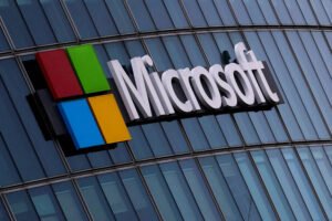 A imagem mostra o logotipo da Microsoft em um edifício de vidro. O logotipo é composto por quatro quadrados coloridos (verde, vermelho, azul e amarelo) dispostos em um formato de grade, ao lado da palavra