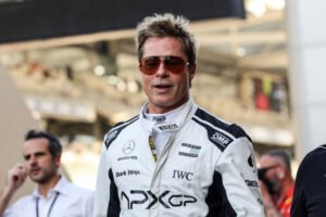 Um homem vestido com um macacão de corrida branco, com detalhes em preto e logotipos de patrocinadores. Ele usa óculos escuros e tem cabelo loiro e desgrenhado. Ao fundo, há pessoas e uma pista de corrida, sugerindo que ele está em um evento de Fórmula 1.