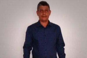 Um homem está posando em pé, com um fundo branco. Ele usa uma camisa azul de manga longa e calças escuras. O homem tem cabelo curto e está sorrindo levemente, com as mãos ao lado do corpo.