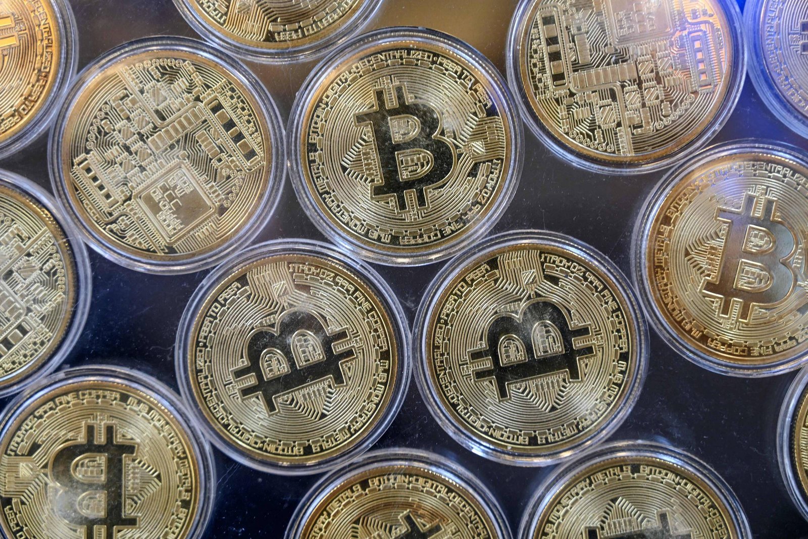 A imagem mostra várias moedas de Bitcoin dispostas em uma superfície. As moedas são de cor dourada e apresentam o símbolo do Bitcoin em destaque, com detalhes de circuitos eletrônicos ao fundo. Elas estão organizadas em um padrão circular, com algumas moedas parcialmente sobrepostas.