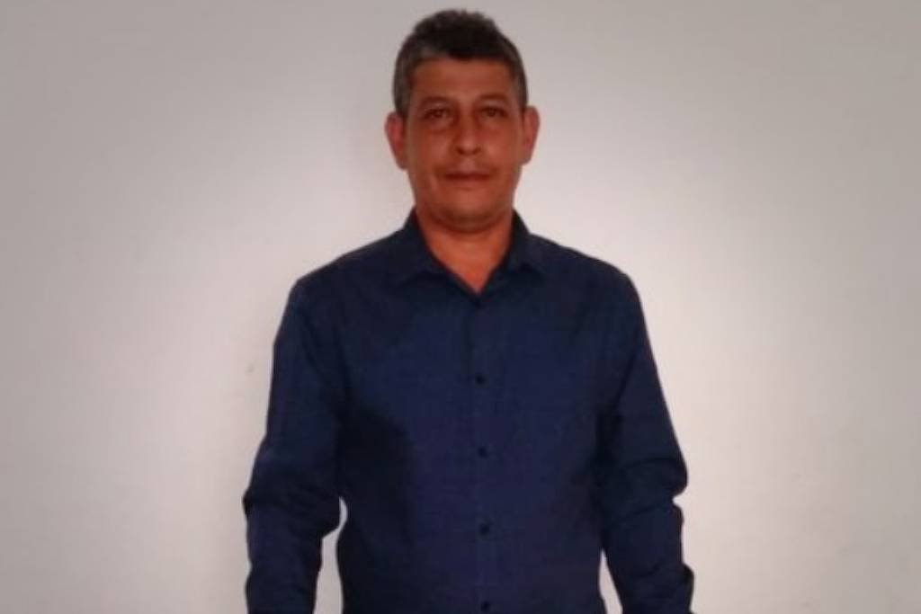 Um homem está posando em pé, com um fundo branco. Ele usa uma camisa azul de manga longa e calças escuras. O homem tem cabelo curto e está sorrindo levemente, com as mãos ao lado do corpo.