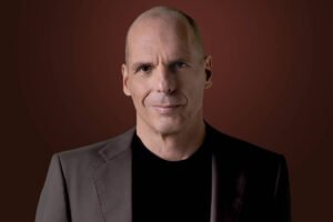 Um homem com cabeça raspada e expressão séria está posando para a foto. Ele usa um terno escuro e uma camiseta preta por baixo. O fundo é de uma cor marrom escura, criando um contraste com sua roupa. O homem parece estar olhando diretamente para a câmera.