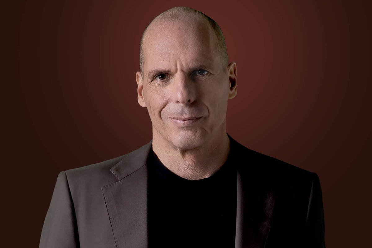 Um homem com cabeça raspada e expressão séria está posando para a foto. Ele usa um terno escuro e uma camiseta preta por baixo. O fundo é de uma cor marrom escura, criando um contraste com sua roupa. O homem parece estar olhando diretamente para a câmera.