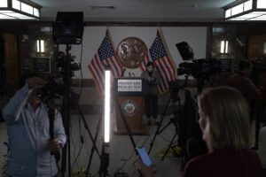 A imagem mostra um cenário de coletiva de imprensa com um púlpito central, onde está escrito