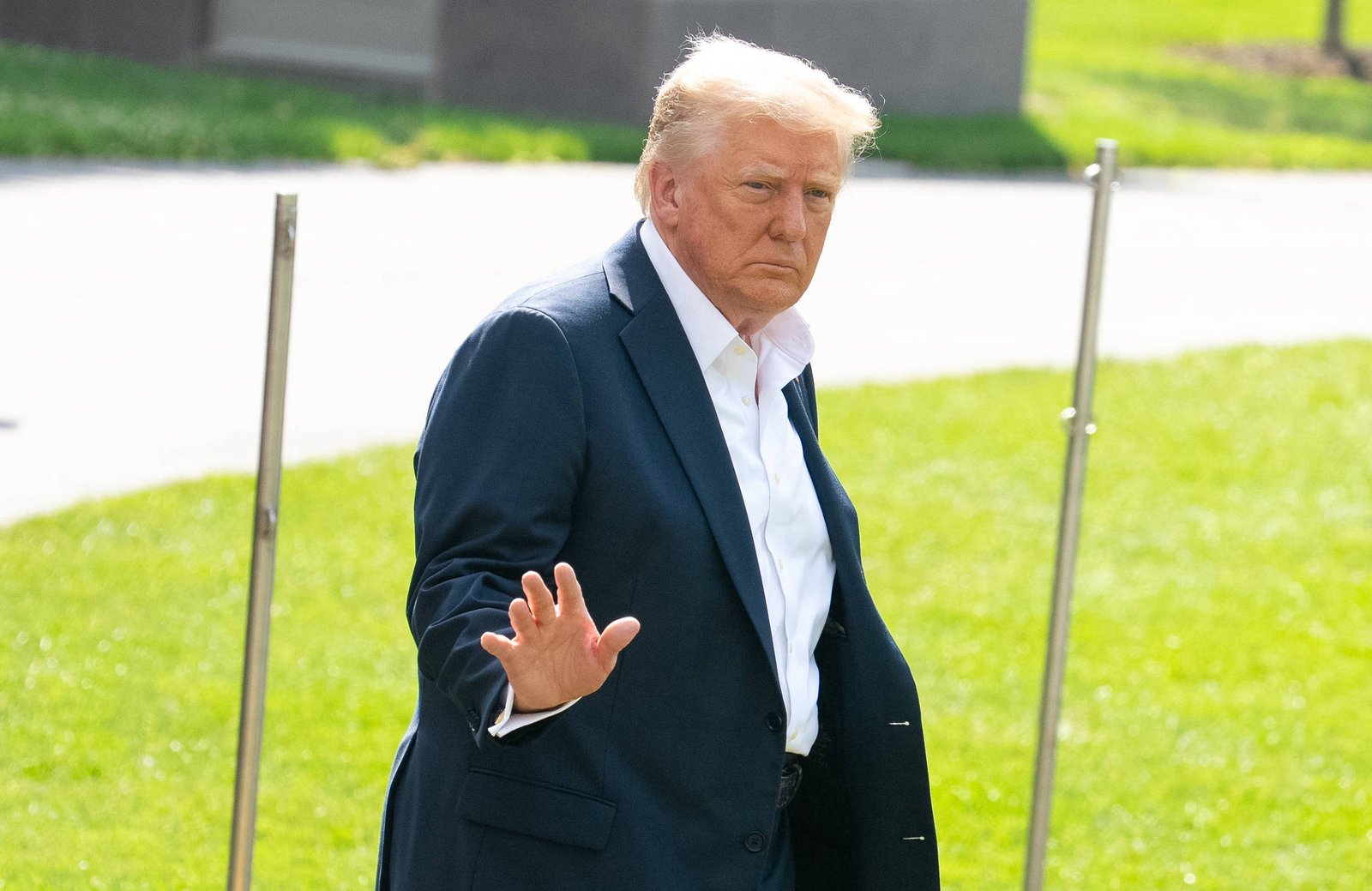 Um homem com cabelo loiro e uma roupa escura está em um ambiente externo, acenando com a mão direita. Ele parece estar em um espaço verde, com grama ao fundo e uma estrutura ao fundo que não é claramente visível. O homem tem uma expressão séria e está olhando para a frente.