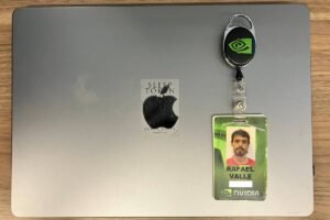 A imagem mostra um laptop da Apple com um logotipo da Apple na parte superior. Ao lado do laptop, há um crachá de identificação com uma foto de um homem e um chaveiro com o logotipo da NVIDIA. O fundo é uma superfície de madeira clara.