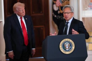 A imagem mostra um evento oficial com duas pessoas. À esquerda está Donald Trump, um homem com cabelo loiro e terno escuro, usando uma gravata vermelha, observa atentamente. À direita está Jensen Huang, um homem de cabelo grisalho, usando óculos e um terno escuro, está falando ao microfone. No fundo, há uma bandeira dos Estados Unidos e o selo presidencial.