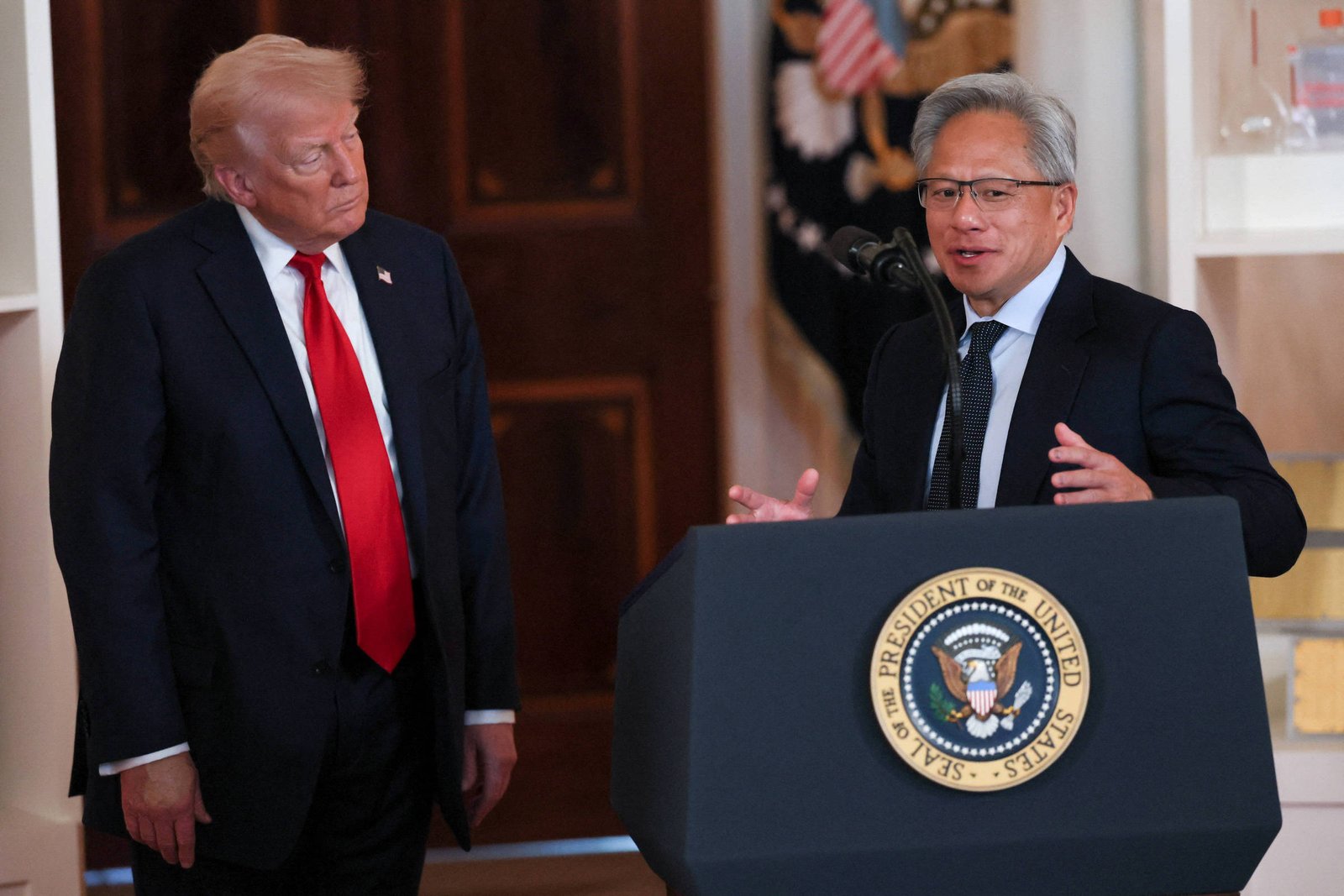 A imagem mostra um evento oficial com duas pessoas. À esquerda está Donald Trump, um homem com cabelo loiro e terno escuro, usando uma gravata vermelha, observa atentamente. À direita está Jensen Huang, um homem de cabelo grisalho, usando óculos e um terno escuro, está falando ao microfone. No fundo, há uma bandeira dos Estados Unidos e o selo presidencial.