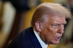 A imagem mostra um homem de perfil, com cabelo loiro e uma expressão séria. Ele está usando um terno escuro e uma gravata amarela. O fundo é desfocado, sugerindo um ambiente formal.