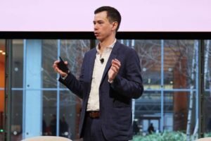 Um homem está em pé em um palco, segurando um dispositivo em uma das mãos e gesticulando com a outra. Ele está vestido com um blazer azul escuro e uma camisa clara. Ao fundo, há uma grande janela com vista para um espaço urbano, com árvores e edifícios visíveis. O ambiente parece ser um auditório ou sala de conferências.