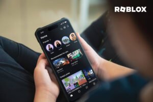 A imagem mostra uma pessoa segurando um smartphone que exibe a interface do aplicativo Roblox. A tela do celular apresenta várias opções de jogos e perfis de usuários. O fundo é desfocado, sugerindo um ambiente interno.
