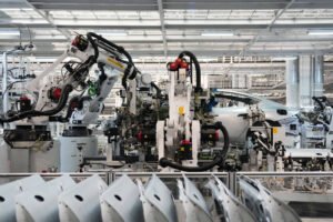 A imagem mostra uma linha de montagem de veículos, com robôs industriais em ação. Vários braços robóticos estão posicionados, manipulando componentes de um carro em um ambiente de fábrica bem iluminado. No primeiro plano, há partes de carrocerias dispostas em uma linha, enquanto ao fundo é possível ver um veículo em processo de montagem.