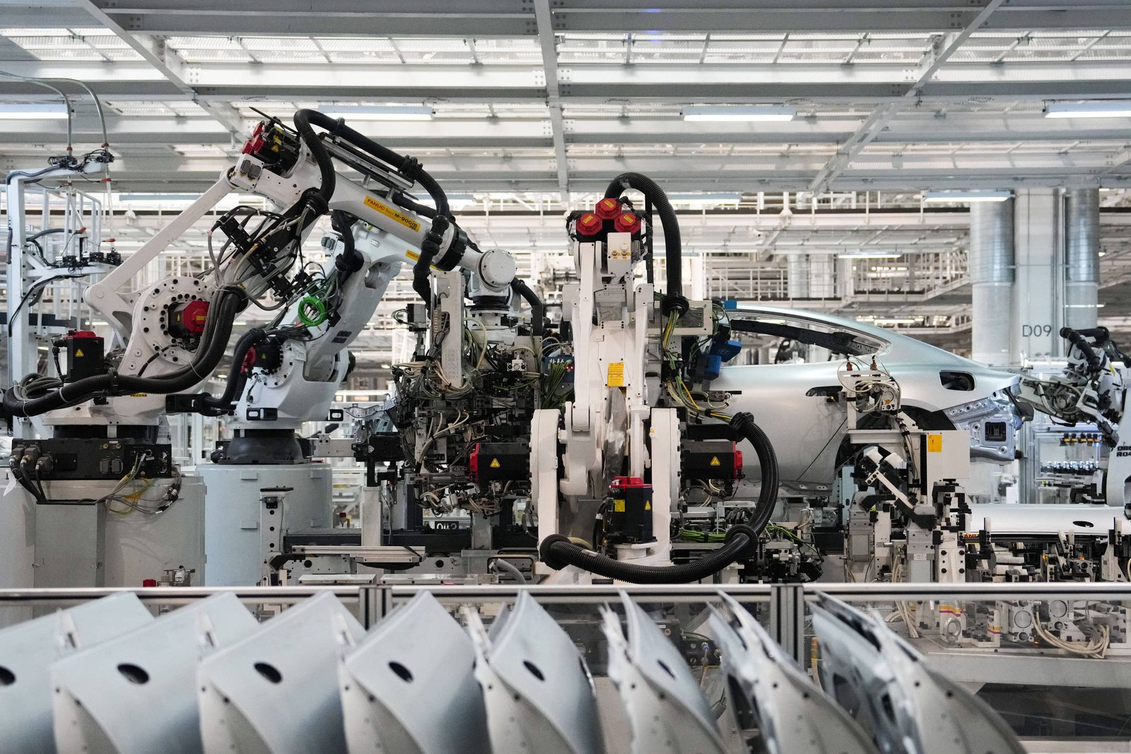 A imagem mostra uma linha de montagem de veículos, com robôs industriais em ação. Vários braços robóticos estão posicionados, manipulando componentes de um carro em um ambiente de fábrica bem iluminado. No primeiro plano, há partes de carrocerias dispostas em uma linha, enquanto ao fundo é possível ver um veículo em processo de montagem.