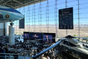 A imagem mostra um evento realizado em um espaço expositivo amplo, com mesas dispostas para os convidados. No fundo, há um grande painel com logotipos de empresas como Palantir, Microsoft e SAIC. À esquerda, um avião presidencial está suspenso, e à direita, um helicóptero. Grandes janelas permitem a entrada de luz natural e oferecem uma vista para uma paisagem montanhosa ao fundo. O ambiente é elegante e moderno, com muitas pessoas presentes.