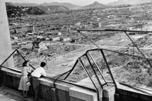 A imagem mostra um casal observando uma vasta área devastada, com muitos edifícios destruídos e terrenos vazios. O cenário é de destruição, com montanhas ao fundo e uma rede de proteção visível na frente. A atmosfera é de tristeza e reflexão sobre a devastação.