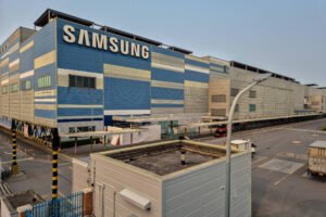A imagem mostra uma grande instalação industrial da Samsung, com uma fachada predominantemente azul e cinza. O edifício é moderno e possui várias janelas. Na frente, há uma área de carga e descarga com caminhões estacionados. O céu está claro, indicando um dia ensolarado.