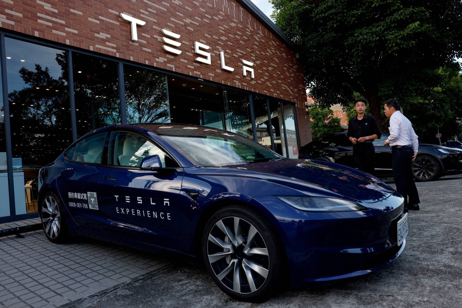 A imagem mostra um carro Tesla de cor azul estacionado em frente a um showroom da marca. O carro possui uma placa com a inscrição