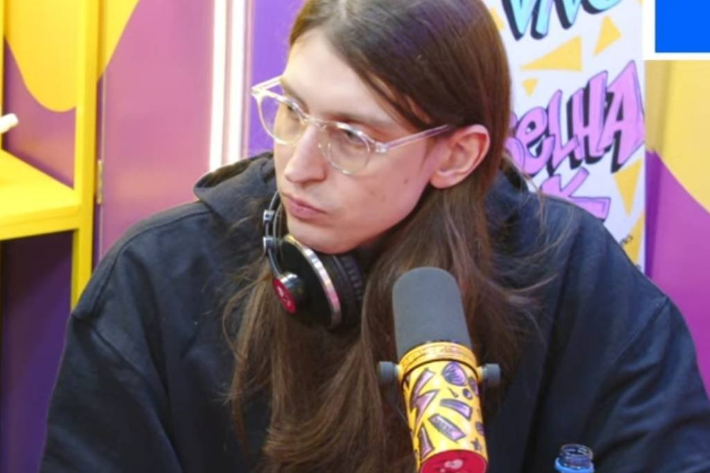 A imagem mostra dois homens sentados em um estúdio colorido. O homem à esquerda tem cabelo escuro e barba, vestindo uma camiseta preta com uma bandeira italiana e texto em português que diz