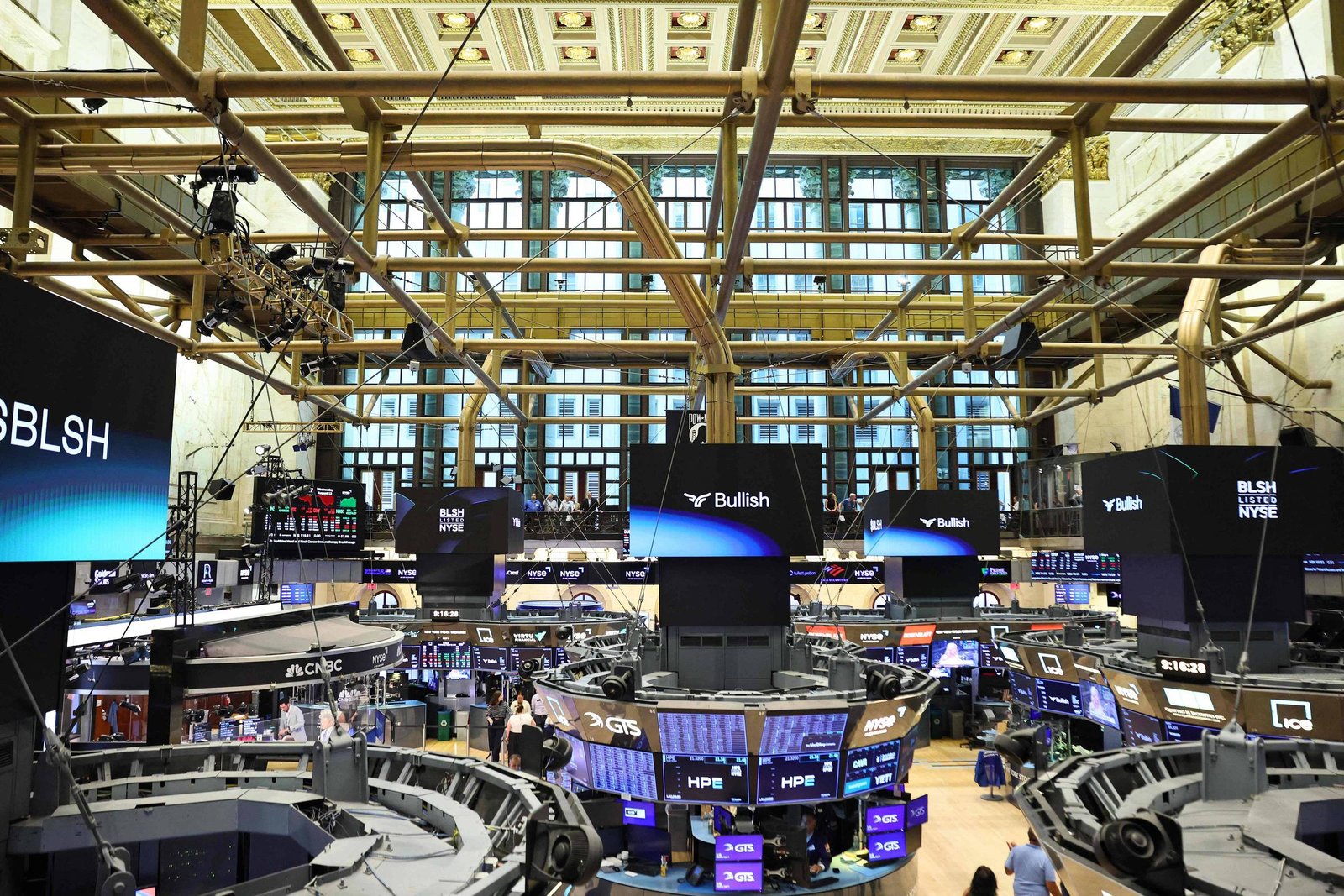 A imagem mostra o interior de uma bolsa de valores, com várias telas exibindo informações financeiras. O ambiente é amplo, com um teto alto e janelas grandes ao fundo. Há mesas de negociação em formato circular e pessoas se movimentando pelo espaço. As telas apresentam dados e gráficos relacionados ao mercado financeiro.