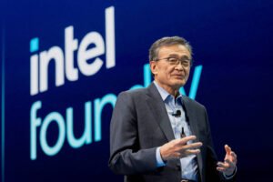 Um homem de óculos e cabelo grisalho está em pé, gesticulando enquanto fala em um palco. Ele usa um paletó escuro e uma camisa clara. Ao fundo, há um grande logotipo da Intel Foundry em letras brancas e azuis.