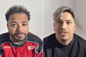 A imagem apresenta dois homens em um fundo neutro. O homem à esquerda está usando um macacão de corrida preto e vermelho, com cabelo encaracolado e uma barba bem cuidada. O homem à direita tem cabelo liso e platinado, com uma barba curta e está vestindo uma camiseta preta. Ambos estão olhando diretamente para a câmera.
