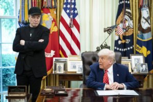 A imagem mostra duas pessoas no Salão Oval da Casa Branca. À esquerda, um homem com um boné preto e roupas escuras está em pé, com uma expressão séria. À direita, um homem de cabelo claro, vestido com um terno escuro e uma gravata vermelha, está sentado à mesa, olhando para o outro. Atrás deles, há cortinas amarelas e bandeiras dos Estados Unidos e do Corpo de Fuzileiros Navais. Na mesa, há alguns objetos e fotos emolduradas.