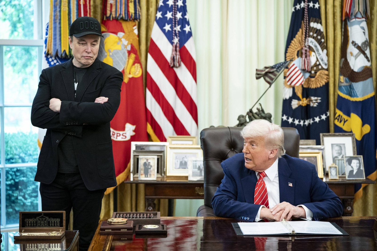 A imagem mostra duas pessoas no Salão Oval da Casa Branca. À esquerda, um homem com um boné preto e roupas escuras está em pé, com uma expressão séria. À direita, um homem de cabelo claro, vestido com um terno escuro e uma gravata vermelha, está sentado à mesa, olhando para o outro. Atrás deles, há cortinas amarelas e bandeiras dos Estados Unidos e do Corpo de Fuzileiros Navais. Na mesa, há alguns objetos e fotos emolduradas.
