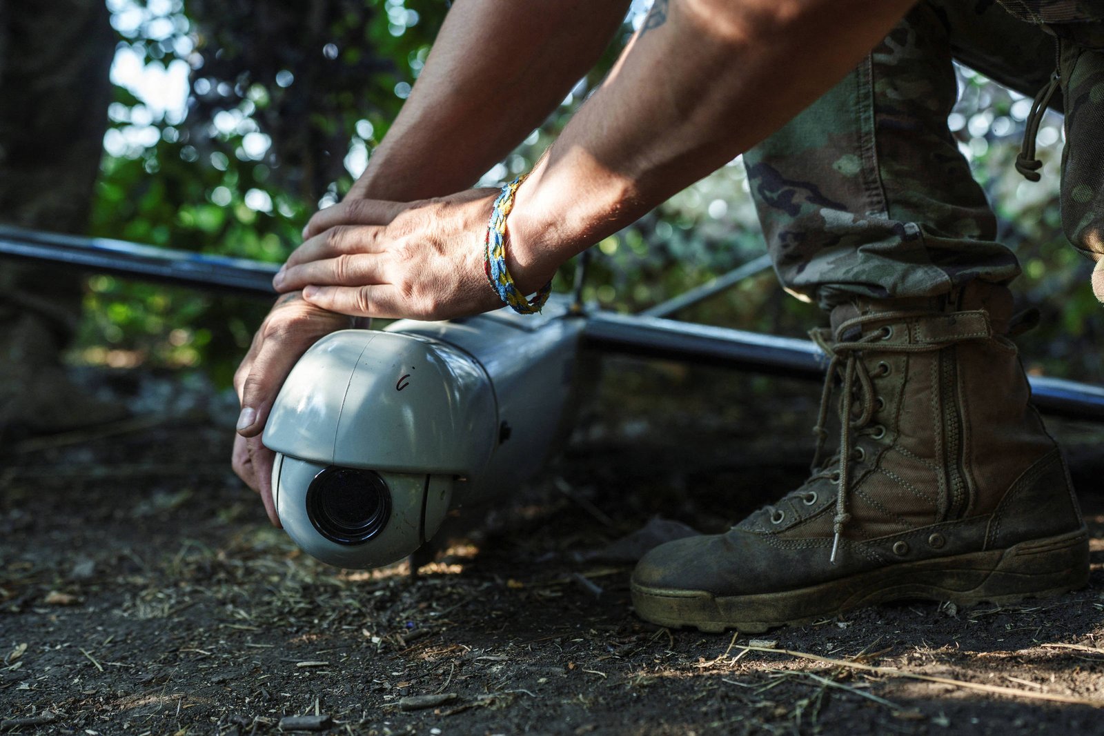 Um soldado em uniforme militar está agachado, segurando um drone com as duas mãos. O drone é de cor cinza e possui uma lente na parte frontal. O soldado está usando botas de combate e uma pulseira colorida no pulso. O ambiente ao redor é natural, com vegetação ao fundo.