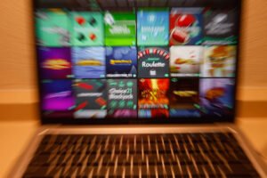 A imagem mostra a tela de um laptop com uma grade de ícones de jogos. Os ícones estão desfocados, mas é possível identificar uma variedade de jogos com diferentes temas e estilos. O teclado do laptop é visível na parte inferior da imagem, e o fundo é de uma superfície clara.