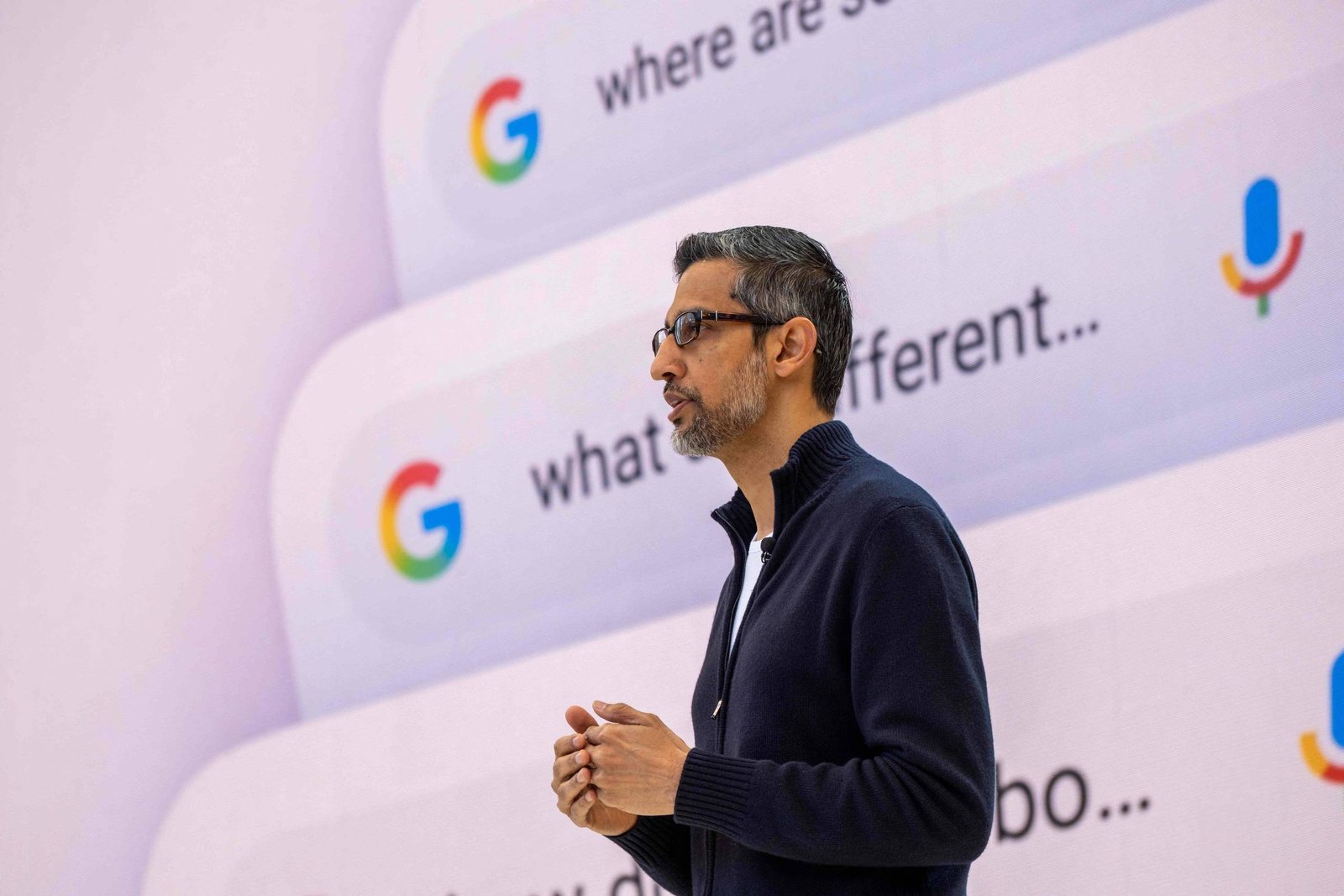 Um homem de cabelo curto e escuro, usando óculos e um suéter escuro, está em pé em um palco, falando. Ao fundo, há uma tela com várias caixas de pesquisa do Google, contendo perguntas como