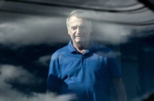 Um homem está em pé, olhando para a câmera, com um sorriso. Ele usa uma camisa azul e está em frente a uma superfície reflexiva que mostra nuvens no céu. O ambiente parece ser ao ar livre, e a luz do sol ilumina a cena.