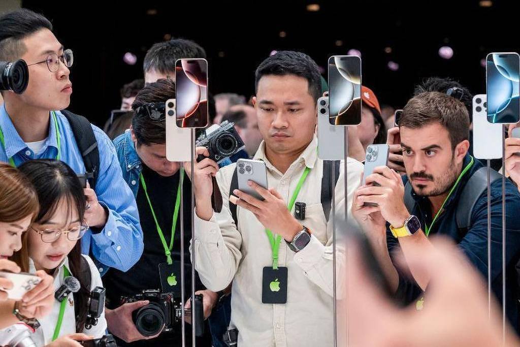 A imagem mostra um grupo de pessoas em um evento, possivelmente de lançamento de produtos, onde várias pessoas estão segurando smartphones e câmeras. Algumas delas usam fones de ouvido e todas parecem concentradas em capturar imagens ou vídeos do que está acontecendo à sua frente. Os participantes estão usando crachás verdes e há dispositivos expostos em suportes ao fundo.