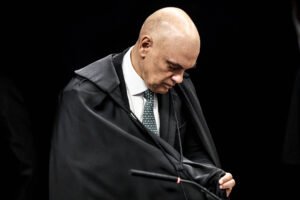 Um homem careca, vestido com um terno escuro e uma gravata, está se curvando levemente para frente enquanto coloca uma capa preta sobre os ombros. O fundo é escuro, sugerindo um ambiente formal, possivelmente uma cerimônia ou evento solene.