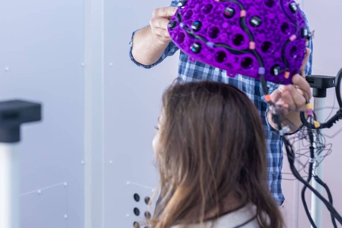 Um homem com cabelo cacheado e óculos está segurando um dispositivo em forma de cérebro, colorido de roxo, enquanto se prepara para colocá-lo na cabeça de uma mulher com cabelo longo e liso. O ambiente é moderno, com paredes brancas e equipamentos tecnológicos visíveis ao fundo.
