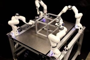 A imagem mostra uma mesa de trabalho com um conjunto de quatro braços robóticos brancos. Os robôs estão posicionados ao redor de uma estrutura cúbica metálica, aparentemente em processo de montagem ou manipulação. A mesa possui uma superfície de metal com iluminação em LED, e o fundo é escuro, destacando os robôs e a estrutura.